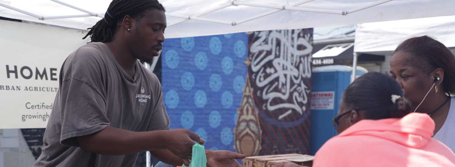 A man in a gray shirt hands items to two women at an outdoor market stall. There is a decorative blue patterned backdrop and a sign that reads Urban Agriculture in the background.